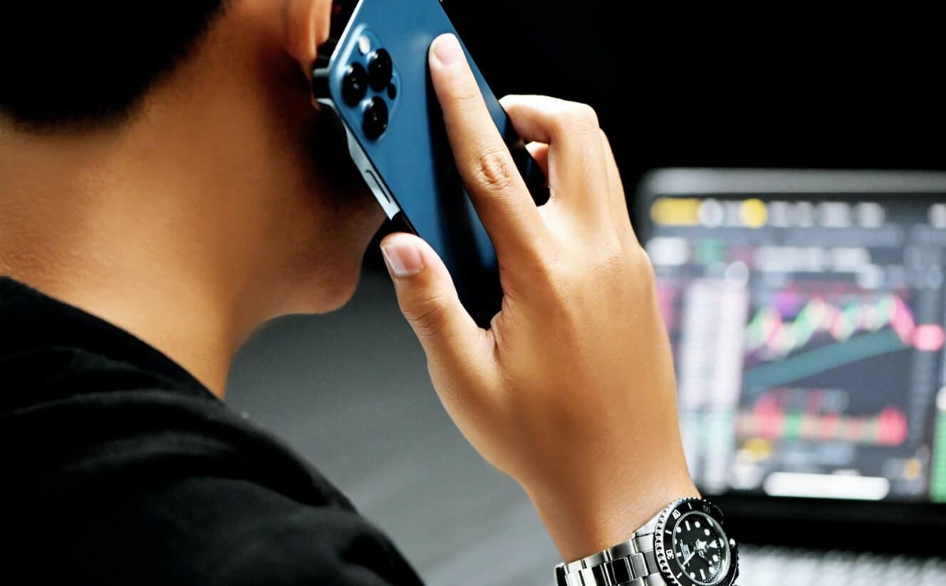 holidng phone to ear with laptop in background showing trading chart